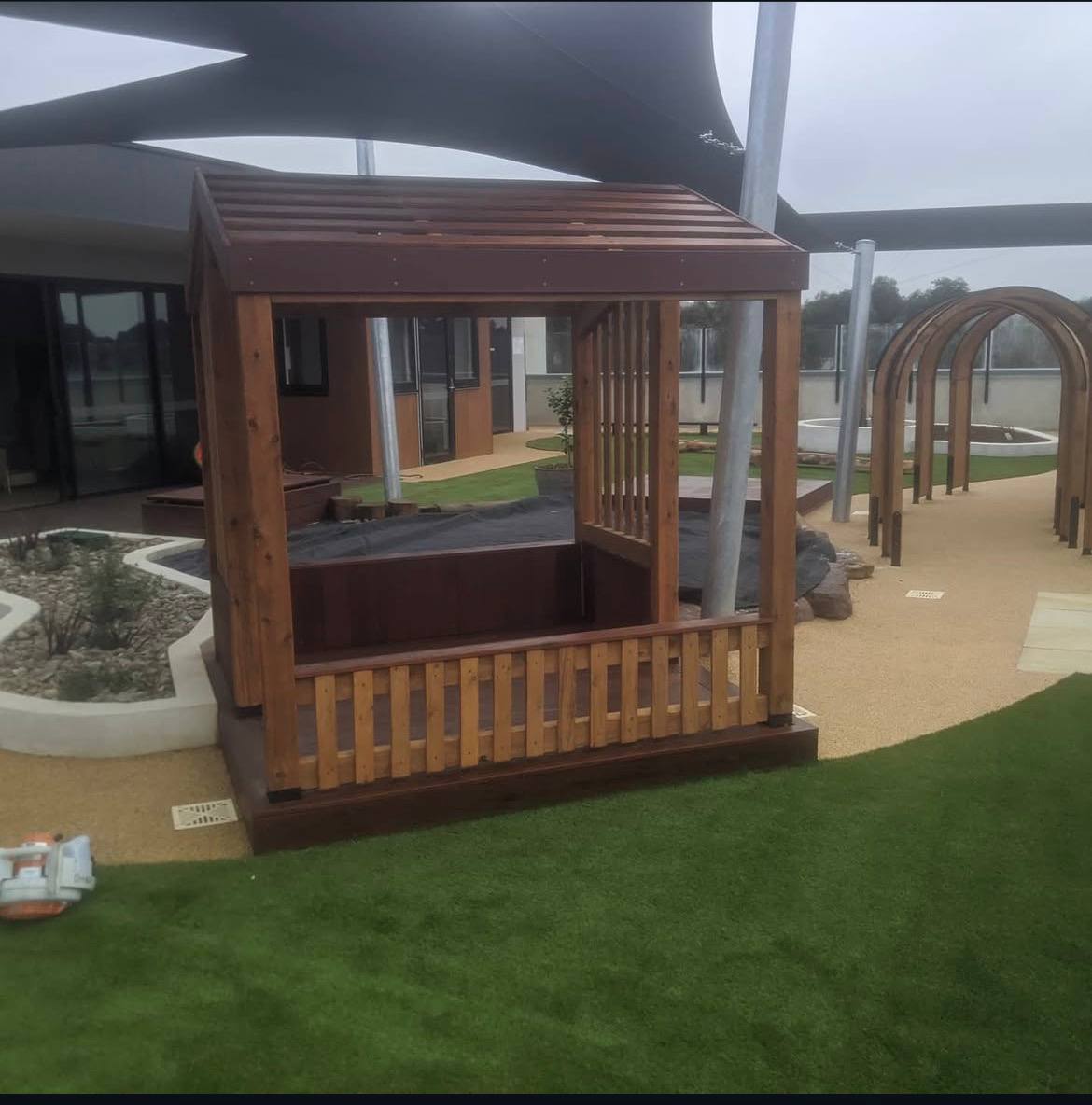 Timber cubby and pergola over a sandpit surrounded by tan wetpour softfall rubber and synthetic turf