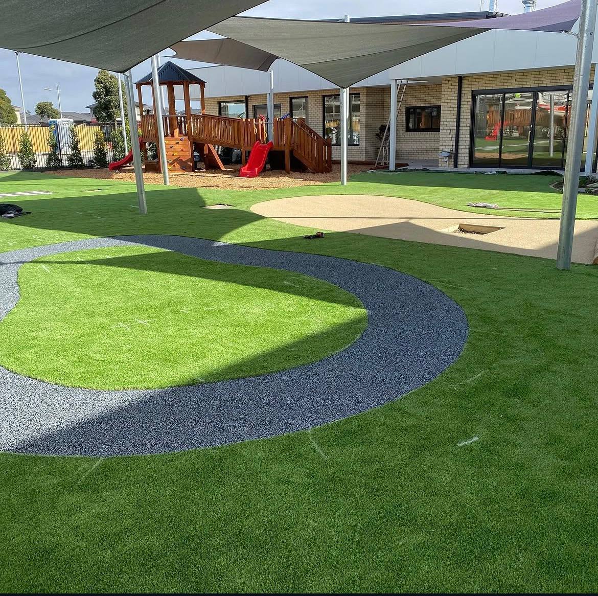 Synthetic turf play area with a curved blue and grey wetpour rubber path leading to a timber playground under shade sails